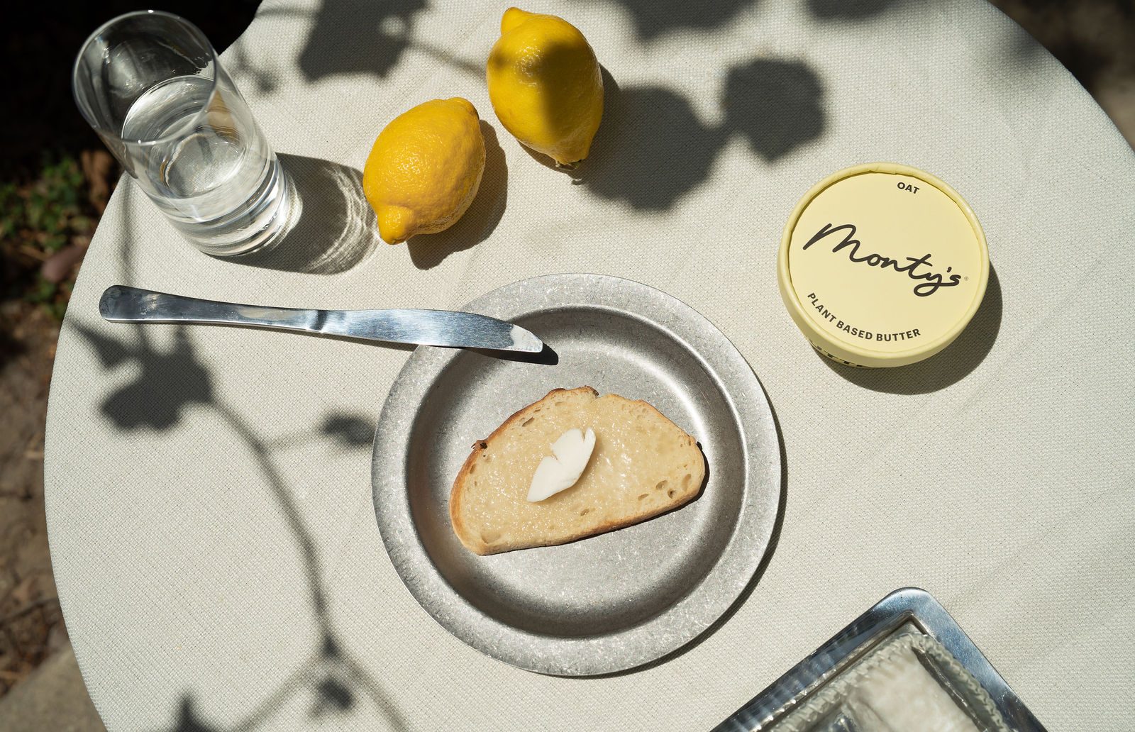 A sunlit garden table set with Monty's Oat Butter, buttered bread, and lemons