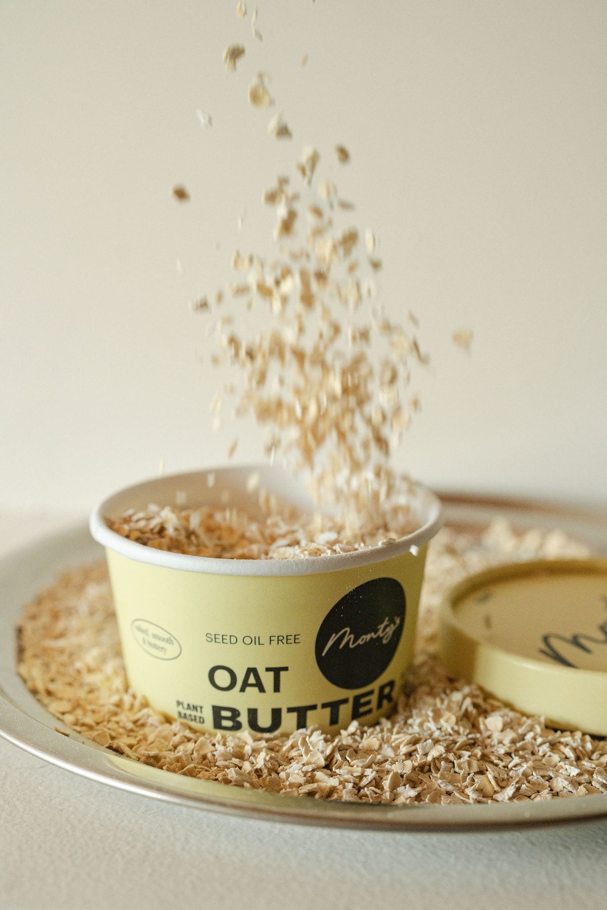 Rolled oats sprinkling into an open cup of Monty's Oat Butter