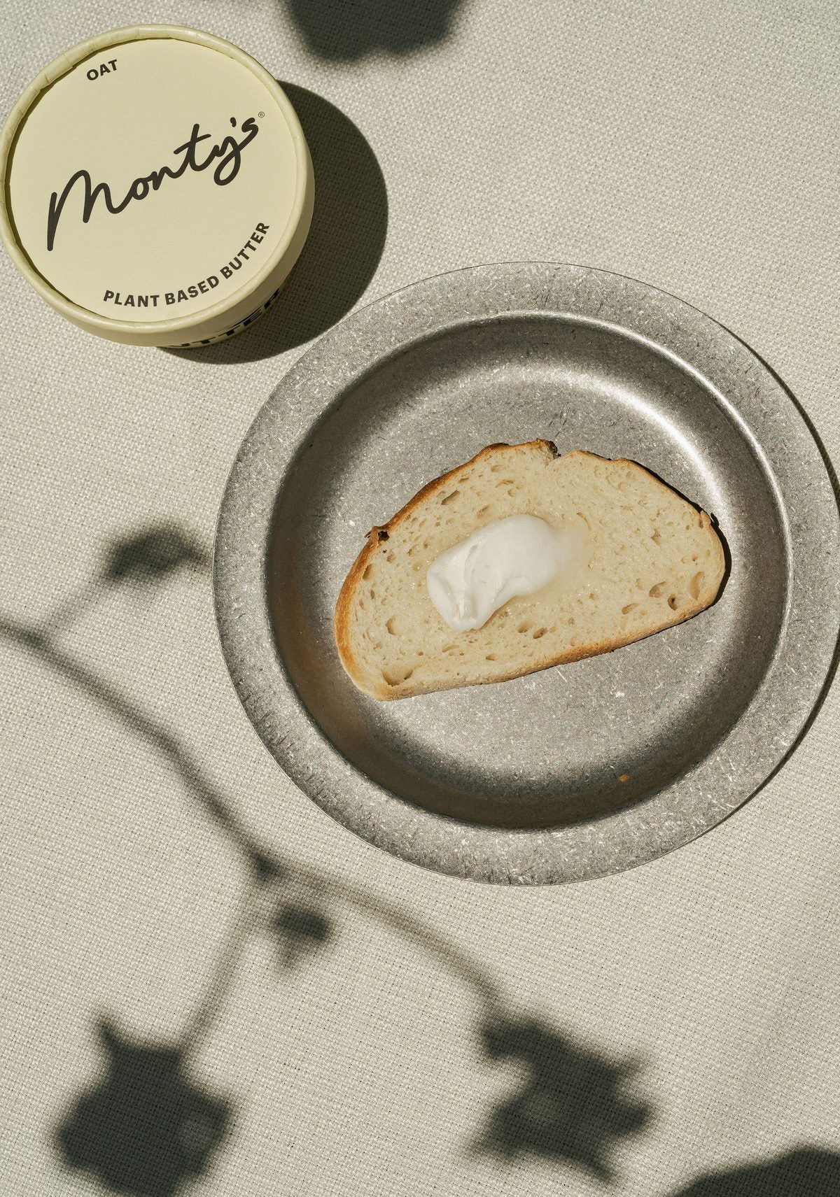 Bread on a pewter plate beside a Monty's Oat Butter cup, with flower shadows